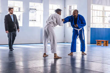 Diverse male athletes wearing gis gripping hands on mats in training studio, coach observing