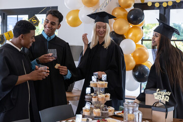 Diverse friends in gowns gathering at table at graduation, sharing cupcakes, toasting wine