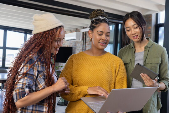 Diverse female coworkers collaborating around laptop at open-plan office desk with tablet - Powered by Adobe