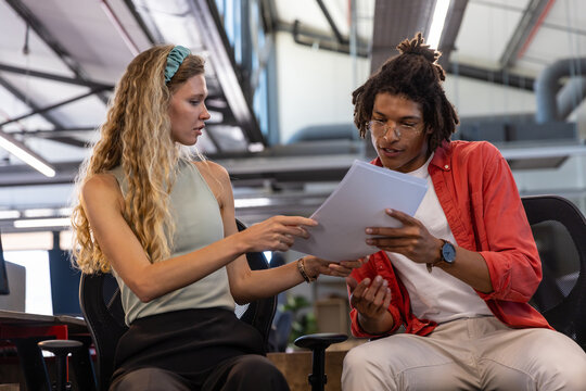 Diverse coworkers reviewing printed documents on swivel chairs by linear lights in open-plan office - Powered by Adobe
