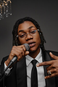 Portrait of young adult Black man wearing glasses holding microphone and gesturing with hand while looking into camera, performing or speaking in formal attire at event