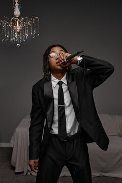 Young adult Black man wearing suit holding microphone performing in indoor setting, standing with confident posture, expressive gesture, chandelier and sofa visible in background