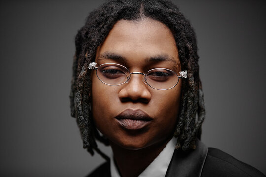 Portrait of young adult Black man wearing eyeglasses looking directly at camera with neutral expression, close up of face and upper shoulders against plain background