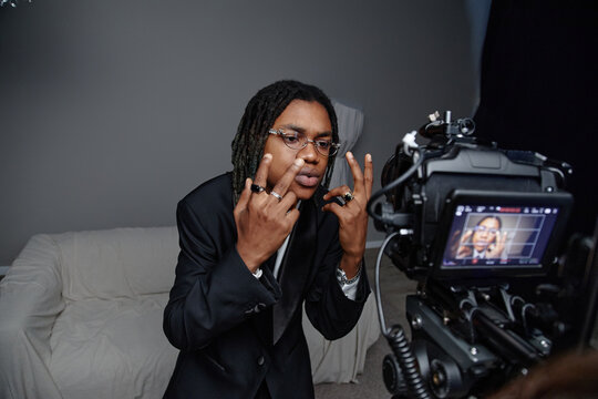 Young adult Black man wearing suit standing in front of professional camera gesturing with both hands and looking directly at lens during video recording session
