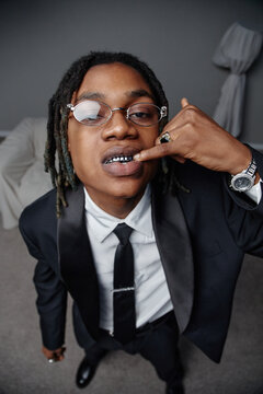 Portrait of young adult Black man wearing suit and tie, standing indoors, looking into camera, showing grillz on teeth with finger, displaying confident facial expression