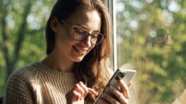 Beautiful young woman wearing glasses smiling while texting on her mobile phone, sitting by a window with her reflection visible and warm sunlight illuminating her face in a cozy home setting