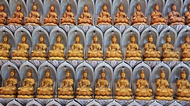 Rows of identical golden Buddha statues in arched niches at Kek Lok Si Temple, Penang. Multiple Guanyin deity sculptures arranged in repetitive pattern on temple wall. Religious art display with