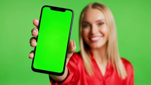 Young Cheerful White blonde woman holding a mobile phone  with green screen, presenting smartphone on chroma key background, for copy-space. Selective focus on phone for technology and app mockup