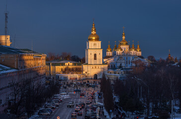 Winter evening view of Sofia Square in Kyiv at sunset, featuring St Michael&rsquo;s Golden Domed Monastery glowing in warm light against deep blue sky and historic city arch