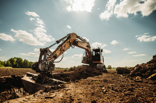 Low angle view of excavator at construction site with dramatic sky. Heavy machinery digging soil and trench for infrastructure works. Industrial earthmoving in rural landscape, sunny day.
