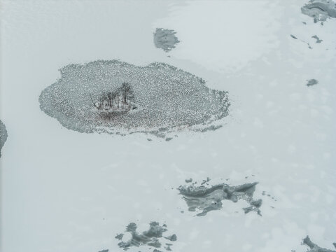 Aerial view of a snow-covered island in Lake Galve with sparse trees, its icy edges dissolving into the vast white expanse, Trakai, Lithuania.