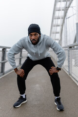Athletic man resting in squat position during outdoor workout on a pedestrian bridge. Focused cardio training, urban fitness routine, strength and endurance exercise, active lifestyle