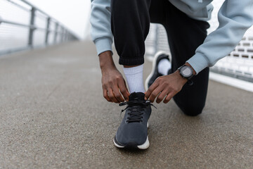Close-up of a focused Black male runner tying shoelaces before training on an urban bridge, sportswear and smartwatch visible, preparation, motivation and healthy active lifestyle