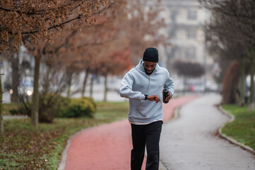 Man in sportswear checking a smartwatch while jogging on a red running track in a foggy park. Outdoor fitness, cardio training, technology in sport, and healthy lifestyle concept