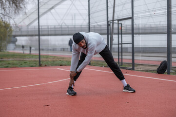 Young athletic man stretching legs during warm-up on an outdoor running track. Focused fitness training, flexibility exercise, urban sports lifestyle and healthy active living.