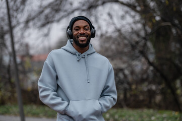 Smiling man in a hoodie and headphones standing outdoors in a park, relaxed after a workout. Active lifestyle, mental wellbeing, outdoor fitness, and positive mood on an autumn day