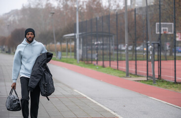 Young Black man wearing a hoodie and backpack waits at a city bus stop on an autumn day, calmly commuting through an urban street, expressing focus, independence, and everyday modern city life