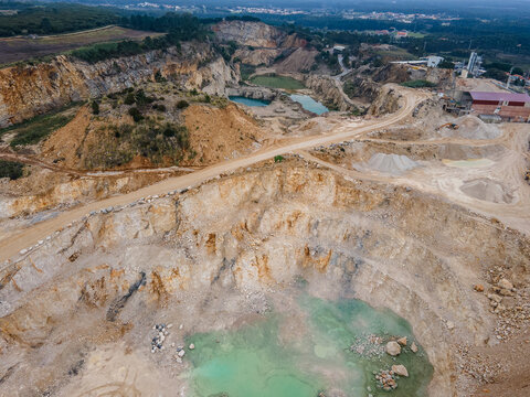 Aerial view of a quarry's stark, layered rock formations meet the pale turquoise water pools, creating a surreal landscape of industry and nature, Foz do Arelho, Leiria District, Portugal.