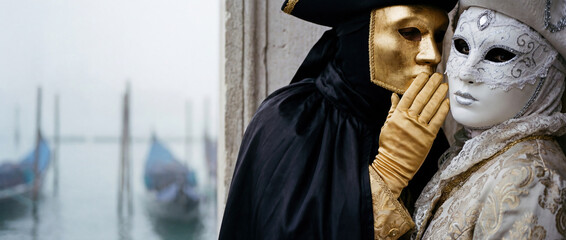 Mysterious couple in Venetian carnival masks whispering secrets by a foggy lagoon. Historical gold and white costumes at the Carnival of Venice with gondolas in the background.