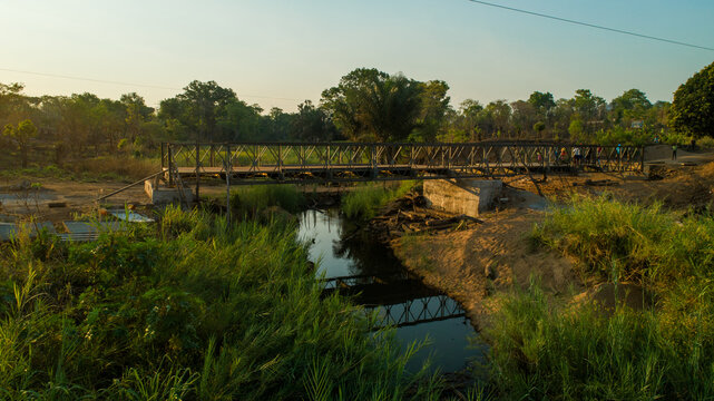 Aerial view of a sturdy steel bridge cutting across a tranquil stream, reflecting the clear sky amid the lush greenery, Derre, Zambezia Province, Mozambique.