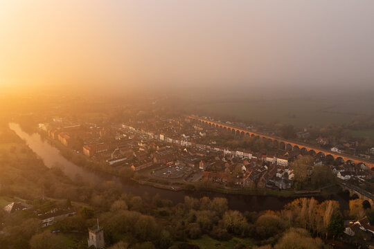 Aerial view of the town bathed in a warm, ethereal glow, the river winding beside clustered buildings and a striking red-brick viaduct stretching across the landscape, Yarm, North Yorkshire, England.