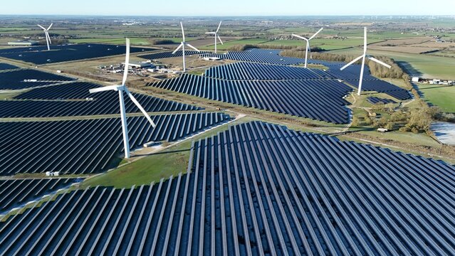 Aerial view of a vast solar farm juxtaposed with towering wind turbines under a clear sky, a symphony of sustainable energy, Wellingborough, England, United Kingdom.