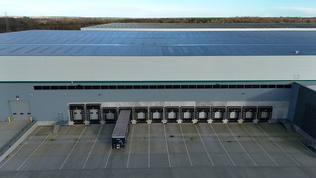 Aerial view of a colossal distribution center with a single truck backed into a loading bay, its dark silhouette a stark contrast against the vast, pale concrete, Kettering, England, United Kingdom.