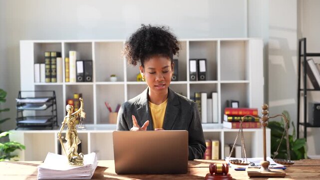 Professional female lawyer offering legal advice and consultation to a client via video conference call, sitting at her desk in a modern law office with a gavel and scales of justice