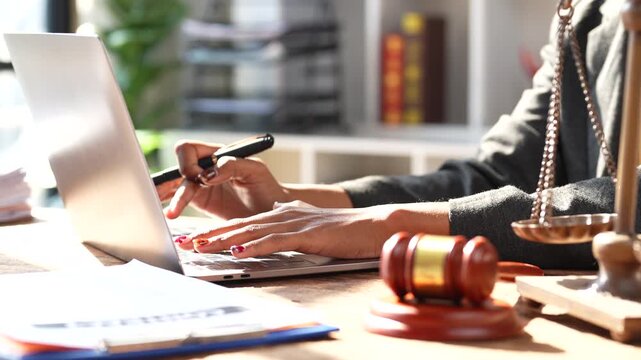 Professional african american female lawyer working on a laptop at her desk with a gavel and scales of justice, focusing on a case, legal advice, and courtroom preparation in a modern law firm