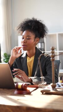 Professional African American female lawyer working at her desk, typing on a laptop and providing online legal consultation to a client while gesturing during a video call in a modern office