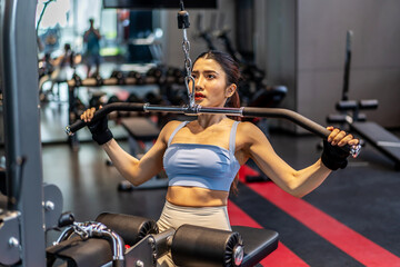 Determined young Asian woman performing wide-grip lat pulldowns on a machine at a modern gym. Professional fitness training, back workout, and healthy lifestyle concept in a luxury health club.