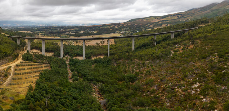 Aerial view of a stark grey bridge cutting through a vibrant green valley, contrasting with the muted tones of the distant mountains, Paarl, Western Cape, South Africa.