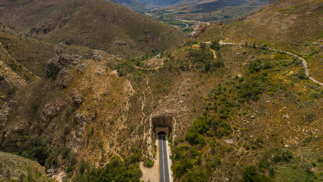 Aerial view of a road disappearing into a dark tunnel carved through rugged mountains, a stark contrast to the bright, sunlit valley beyond, Paarl, Western Cape, South Africa.