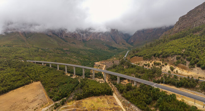 Aerial view of a long bridge cutting through lush green valleys and towering, cloud-kissed mountains, Paarl, Western Cape, South Africa.