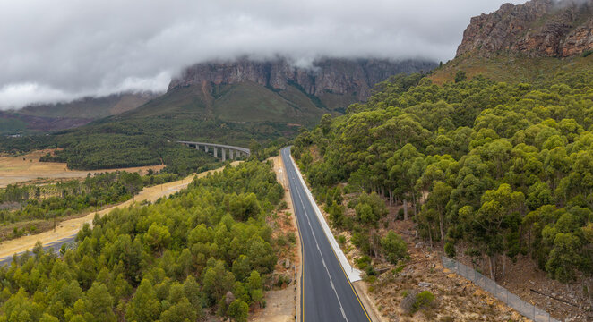 Aerial view of a stark road cuts through lush green forests towards a distant viaduct and cloud-shrouded mountains, Paarl, Western Cape, South Africa.
