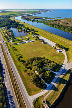 Aerial view of a vibrant green park contrasts with the flowing river and the adjacent highway, creating a dynamic landscape, Lake Okeechobee, John Stretch Memorial Park, Florida, United States.