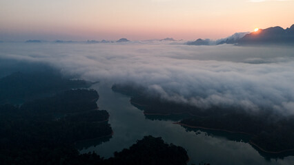 Aerial view of a serene lake embraced by rolling mist and dramatic limestone peaks under a pastel sky at dawn, Khao Sok National Park, Surat Thani, Thailand.