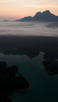 Aerial view of turquoise waters snaking through lush, dark forests beneath a blanket of ethereal mist, crowned by distant, jagged mountains, Khao Sok National Park, Surat Thani, Thailand.