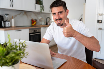Young man smiling at the camera and giving a thumbs up gesture. Working remotely from home on a laptop in a modern kitchen setting
