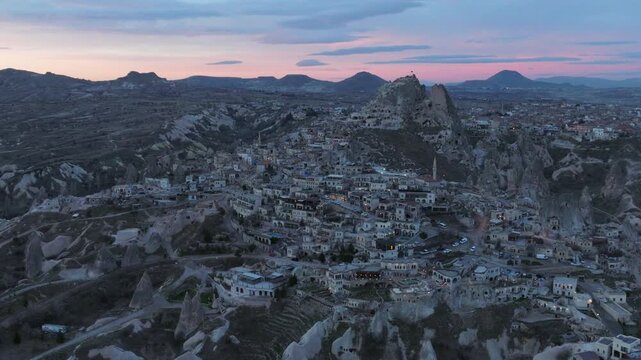 Aerial drone footage of U&ccedil;hisar village in Cappadocia, Turkey, dominated by the iconic U&ccedil;hisar Castle rising above stone houses and cave dwellings. The scene captures the town built into volcanic rock