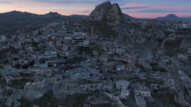 Aerial drone footage of U&ccedil;hisar village in Cappadocia, Turkey, dominated by the iconic U&ccedil;hisar Castle rising above stone houses and cave dwellings. The scene captures the town built into volcanic rock
