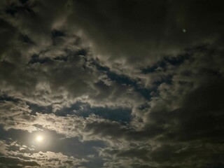 A bright full moon in a cloudy night sky. This phenomenon highlights the reflected sunlight from the moon against the dark sky, while the cloud cover softens it.