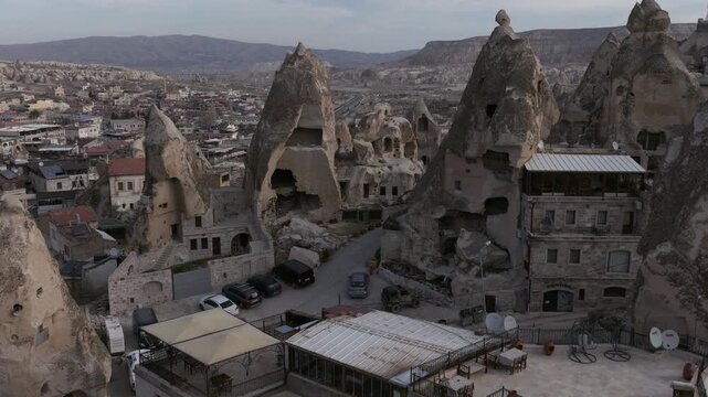 Aerial drone footage of G&ouml;reme town in Cappadocia, Turkey, surrounded by fairy chimneys and volcanic rock formations. The shot reveals traditional stone houses, cave dwellings carved into soft tuff ro