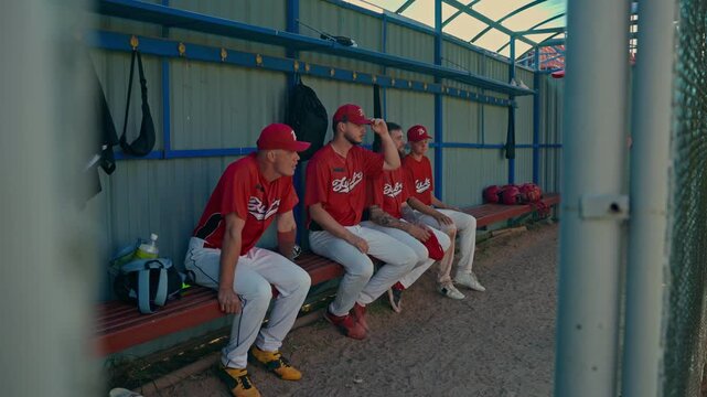 The players sit on a bench in the locker room and listen to the coach.. The baseball team sits and chats on the grass.