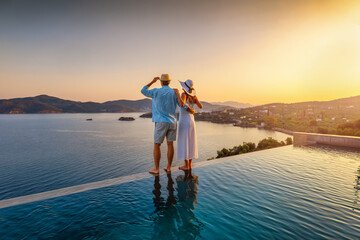 A romantic couple on summer vacation stands by the infinity swimming pool enjoys a the sunset view behind the ocean