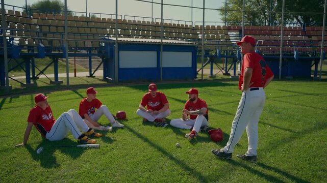 The coach talks to the baseball team after the game. The baseball team sits and chats on the grass.