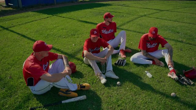 Baseball players laugh and talk after the game. The baseball team sits and chats on the grass.