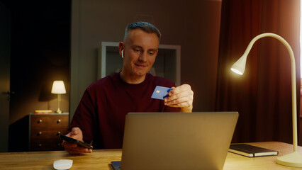 Young adult man sit desk home pay by bank credit card buy stuff online use laptop computer, mobile cell phone. Smiling person do purchase internet store. Remote shopping. Big holiday sale, discounts.