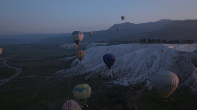 Aerial drone footage of colorful hot air balloons flying above the white travertine terraces of Pamukkale, Turkey, at sunrise. The balloons float over the mineral formations and thermal pools, creatin