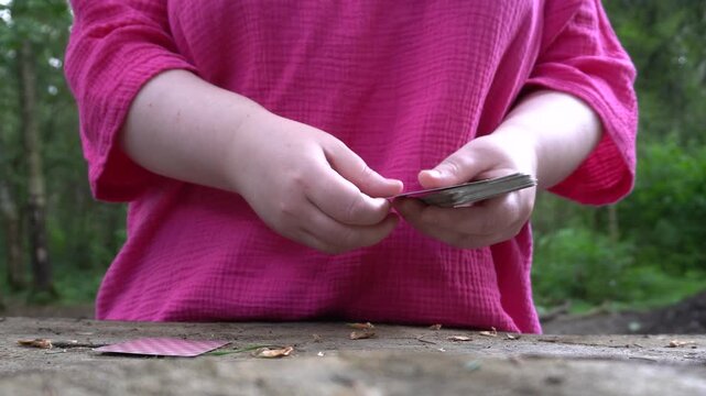 Hands lay out a solitaire card game on a tree stump in the forest. Deck with shirt, close-up, street photography.